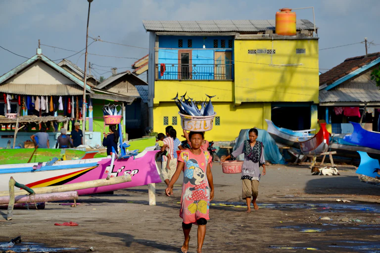 A woman balancing a basket of caught fish on her head, with a yellow house in the background.