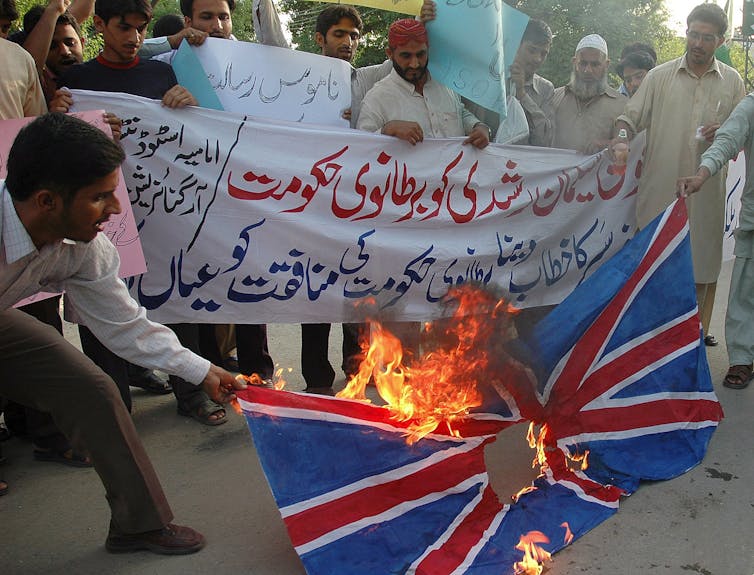 Demonstrators burn a UK flag and carry a banner with Arabic text in black and red.