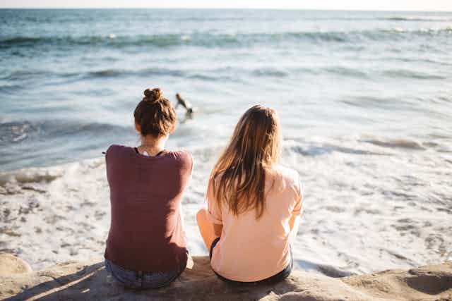Two women sit side by side on a rock looking out at the ocean