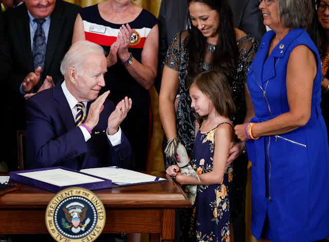 A young girl stands next to a desk where a middle aged white man dressed in a business suit claps his hands after signing a document honoring her father.