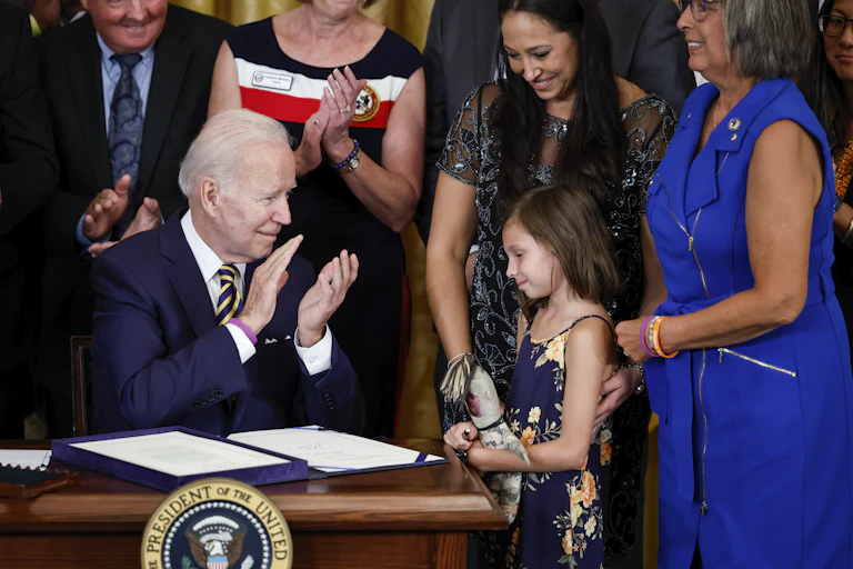 A young girl stands next to a desk where a middle aged white man dressed in a business suit claps his hands after signing a document honoring her father.