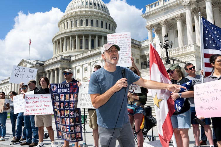 A  man wearing a baseball cap and t-shirt holds a microphone and speaks to a crowd holding flags and waving posters.