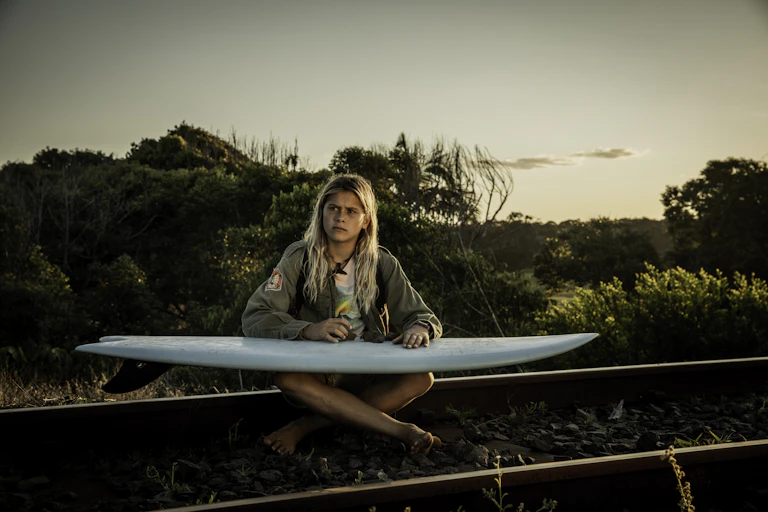 A teenage boy sits with a surfboard