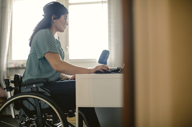 Women with dark hair past her shoulders sitting at her desk. She is wearing a green poke dot top and black pants. She is sitting in a wheel chair with headphones in her ear as she works from her computer. 
Is NDIS Fraud on the rise? 