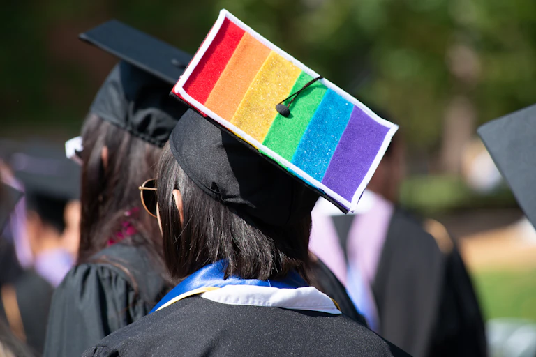 A young woman wears a mortarboard graduation cap emblazoned with the rainbow colors of the gay pride flag.