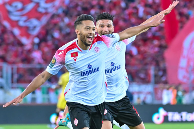 Two men in football jerseys in a stadium run with their arms out like aeroplane wings and big smiles on their open mouths.