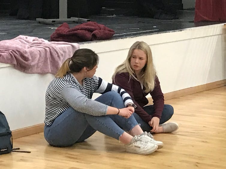Two teenage girls sit on floor and talk to each other.