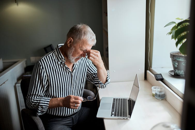A senior man sitting at his computer pinches the roof of his nose, indicating a headache.