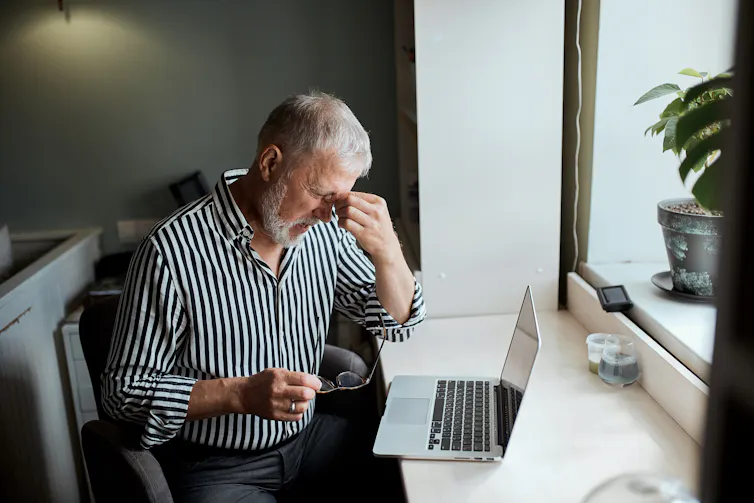 A senior man sitting at his computer pinches the roof of his nose, indicating a headache.