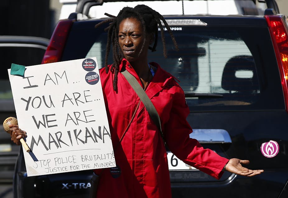 A woman wearing a red work overall stands in front of a car, holding a handwritten poster