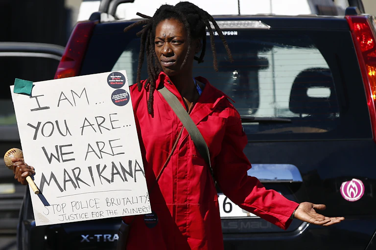 A woman wearing a red work overall stands in front of a car, holding a handwritten poster