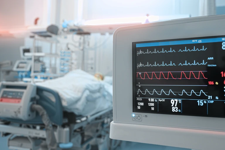 Monitor de constantes vitales y cama con enfermo al fondo.