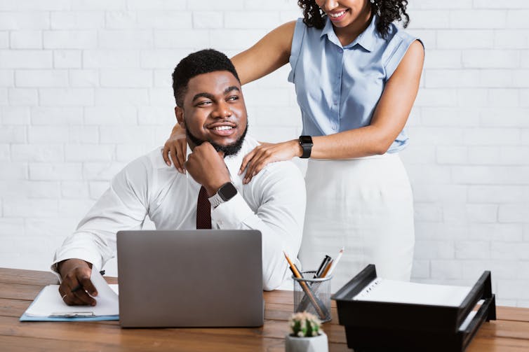 A man sits at his desk while a female colleague touches his shoulders.