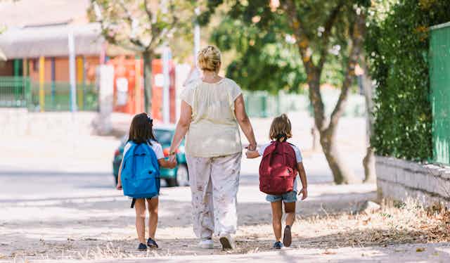 A woman walks toward a school holding hands with two children wearing backpacks.