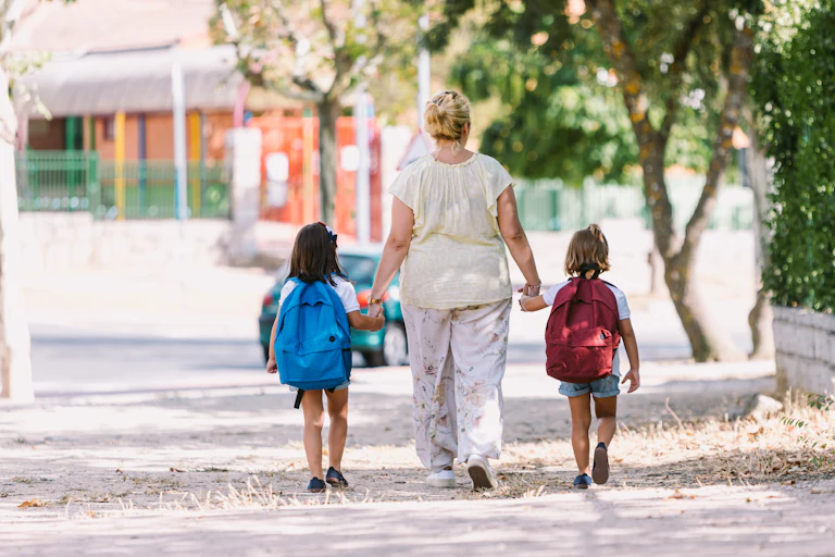 A woman walks toward a school holding hands with two children wearing backpacks.