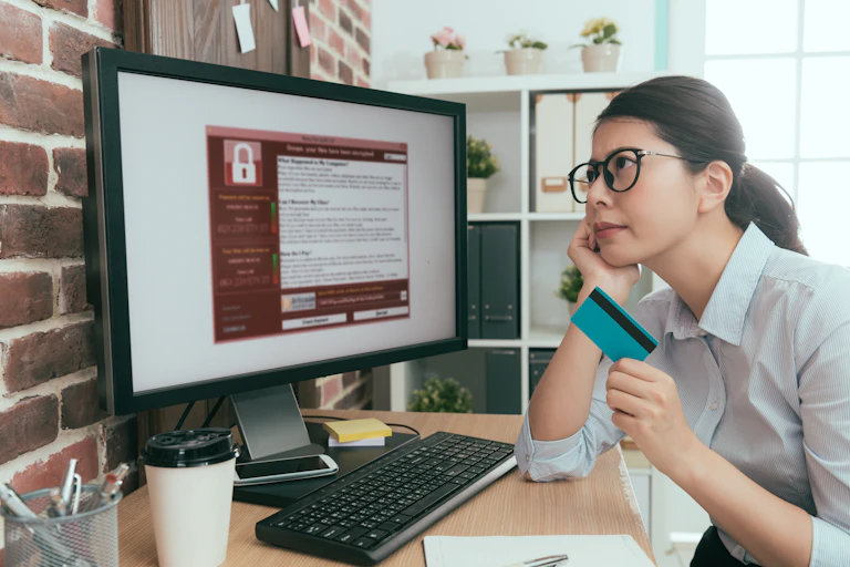 a woman holding a credit card looks at a computer screen with a red warning box showing a padlock icon