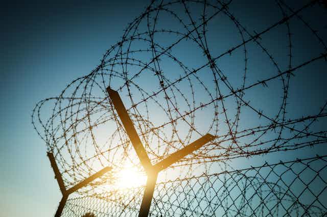 Loops of barbed wire top a metal fence with the sun shining behind it