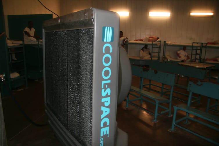 A large vertical fan blows air toward incarcerated people on bunk beds