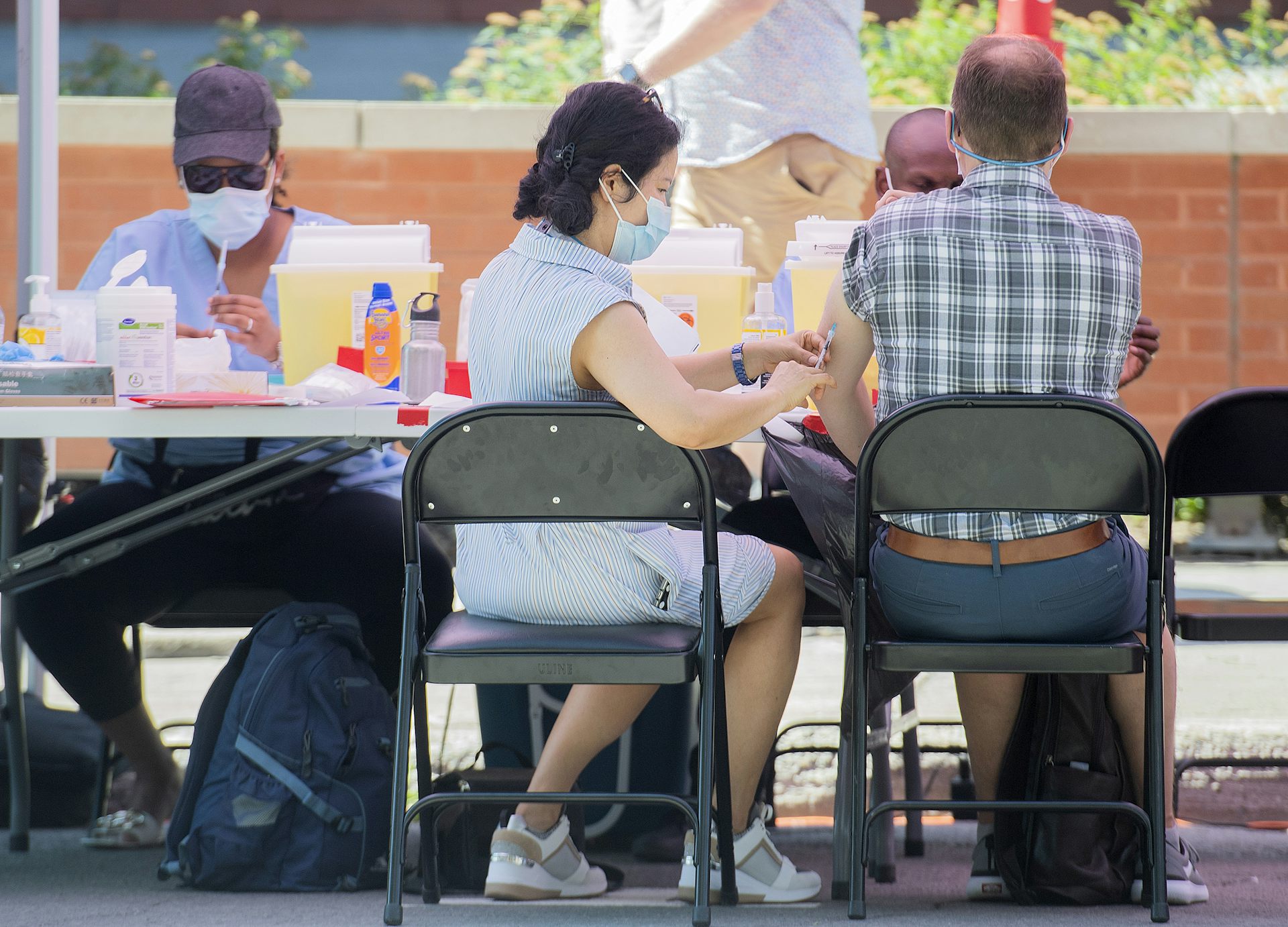 A man in a plaid shirt getting an injection from a woman in a blue dress and face mask, both sitting in folding chairs, seen from behind
