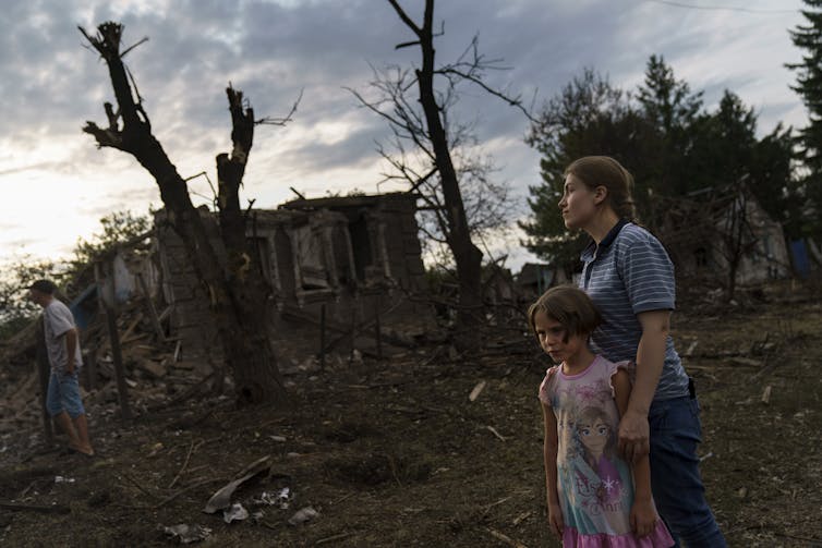 A young woman and a girl stand together amid destroyed homes, looking sad.