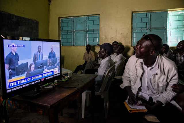 Men sit in a room with yellow walls and green window shades, watching trial proceedings on TV.