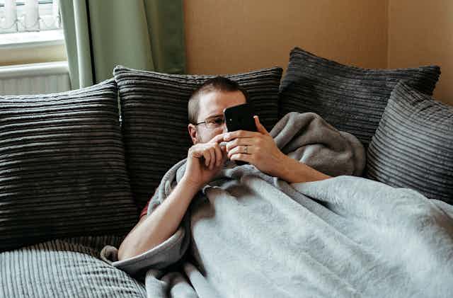 Man on couch under a blanket looks at phone