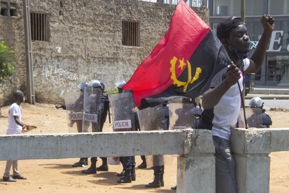 A young male protester with his left fist raised carries the Angolan flag while another chats to police armed with shields at a barricade.