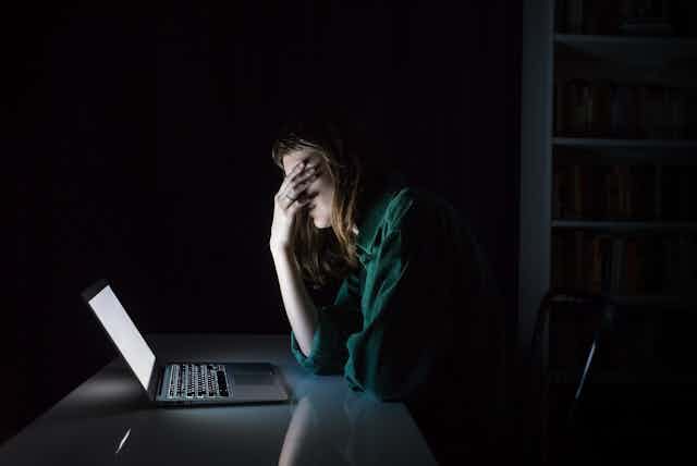 A woman sits in the dark with her head in her hands, in front of a glowing laptop screen