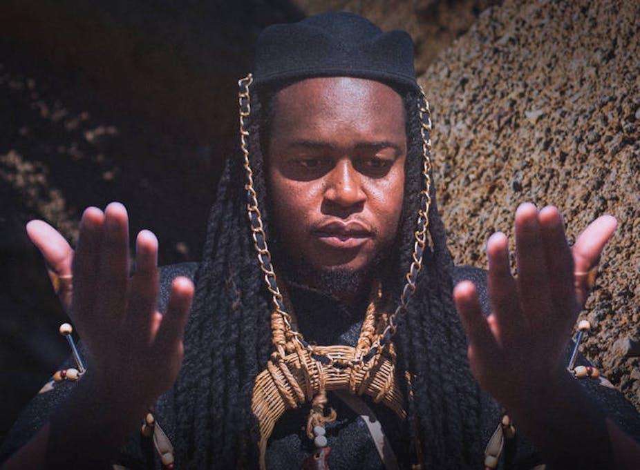 A man in traditional attire and dreadlocks holds up his hands as if in praise or prayer.