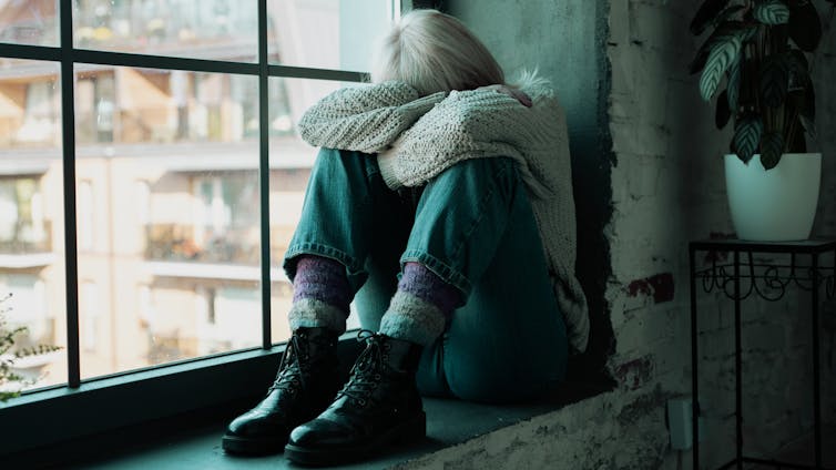 A woman sits by a window, hiding her head.