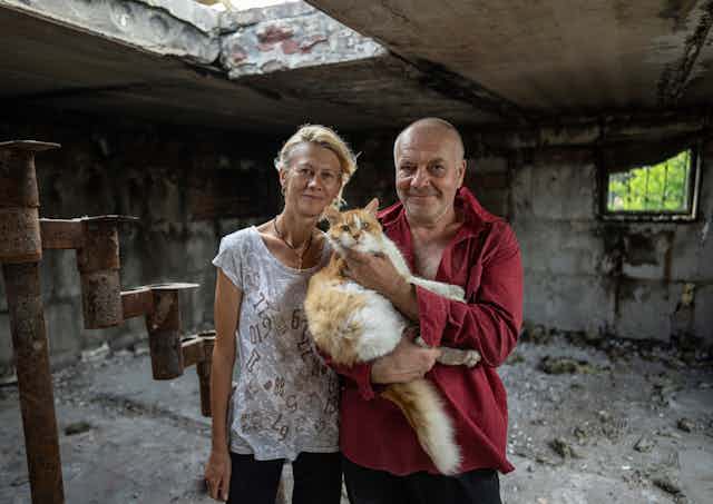 A blond woman in a white shirt and a bald man in a red shirt hold a fluffy cat in the ruins of a bombed house.