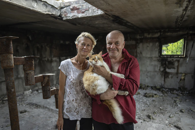 A blond woman in a white shirt and a bald man in a red shirt hold a fluffy cat in the ruins of a bombed house.