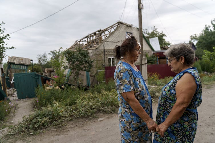 Two older women in blue dresses hold hands outside a destroyed home.
