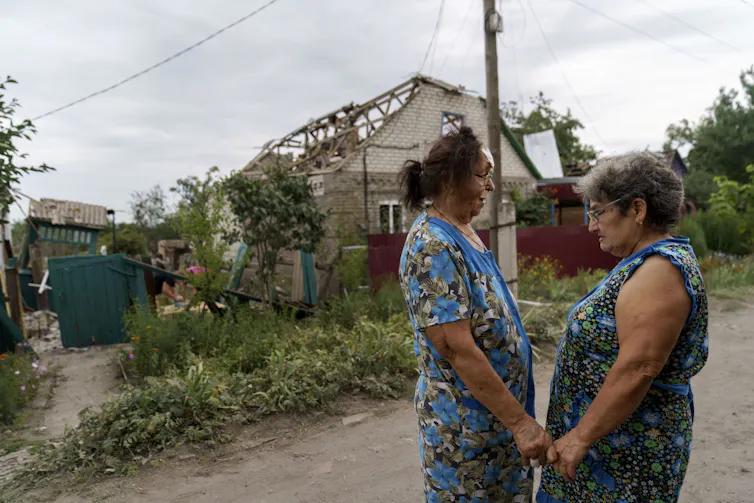 Two older women in blue dresses hold hands outside a destroyed home.
