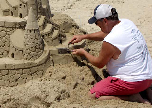 a man in a sleeveless shirt, shorts and hat uses a pair of small trowels to work on a large sandcastle