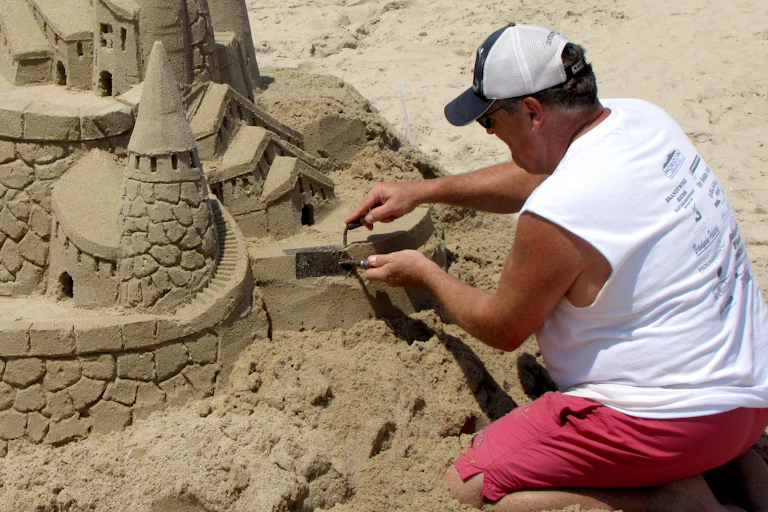 a man in a sleeveless shirt, shorts and hat uses a pair of small trowels to work on a large sandcastle