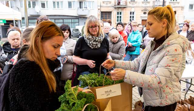 Women hand out paper bags of kale at an open air market.