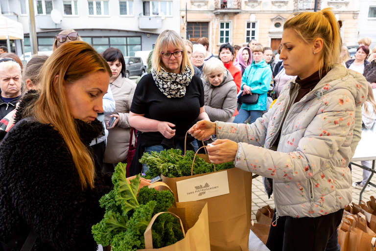 Women hand out paper bags of kale at an open air market.