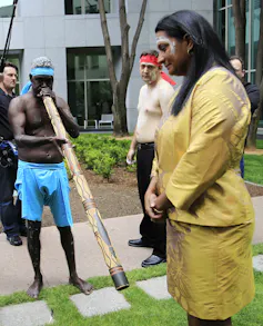 Nova Peris stands with ochre on her forehead as a person plays the didjeridoo beside her. This is ceremony celebrating her swearing into parliament.