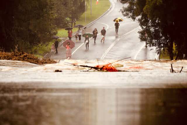 people with umbrellas stand next to flooded road