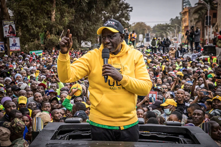 Man speaking into a microphone to address a crowd from the rooftop of a car.