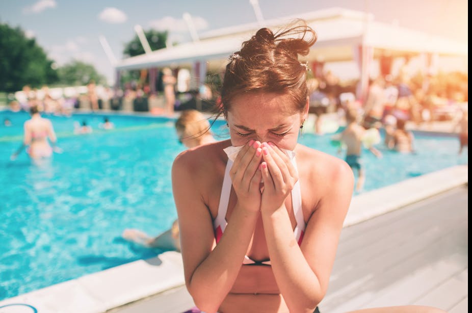A woman sitting near a busy outdoor pool in the summer blows her nose into a tissue.