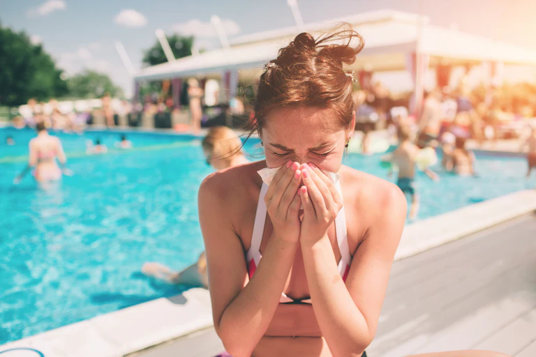 A woman sitting near a busy outdoor pool in the summer blows her nose into a tissue.
