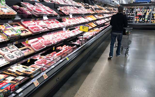 A man pushes a cart along side of an open aisle of meats in a grocery store