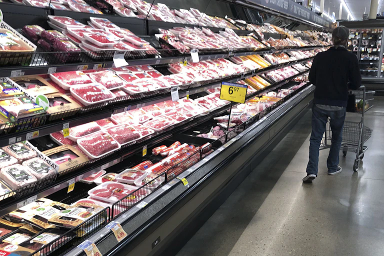A man pushes a cart along side of an open aisle of meats in a grocery store