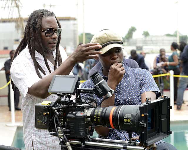 A film set, cordoned off. A dreadlocked man in shades points his hand, talking with a younger man in a cap behind a camera.