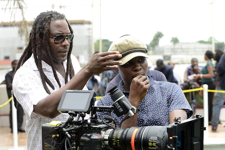 A film set, cordoned off. A dreadlocked man in shades points his hand, talking with a younger man in a cap behind a camera.