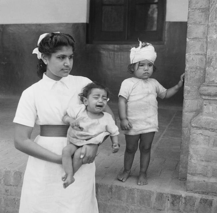 A nurse holds one child while another child with a bandaged head stands next to her.