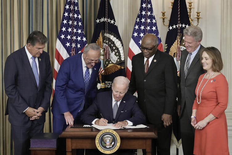 Biden sits at a desk signing the legislation. Sens. Joe Manchin (D-WV.) and Chuck Schumer (D-NY) and Reps. James Clyburn (D-SC), Rep. Frank Pallone (D-NJ) and Rep. Kathy Catsor (D-FL) look over his shoulder.