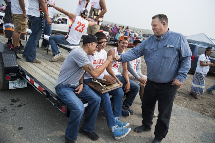 A large man in a blue shirt fistbumps a bunch of young men sitting on a flatbed truck at a rural gathering.